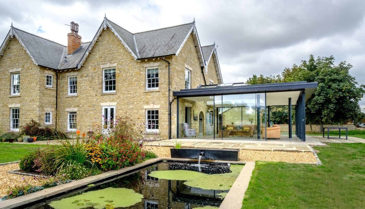 a glass extension to a farm house shown from outside viewed across a pond and green lawn