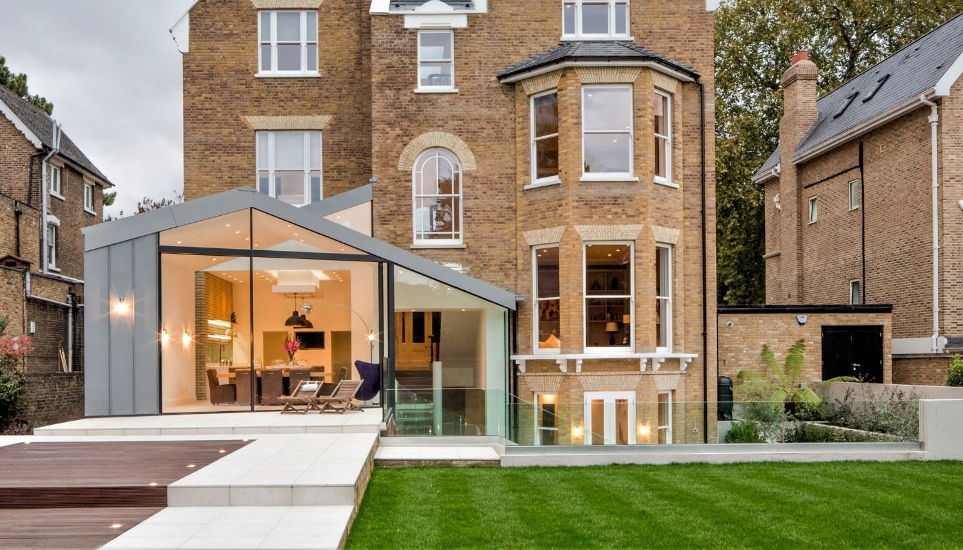 a modern glass extension with a pitched roof shown at dusk with lights on inside