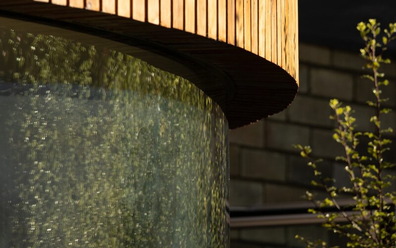 a close up image of the outside of a curved frameless window showing the connecting between the curved glass and the timber cladding