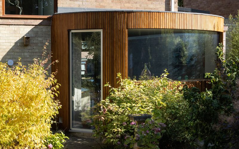 An external view of a curved, timber clad house extension with a frameless curved window