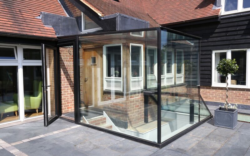 a glass box over a staircase leading to a basement set on a grey patio attached to a traditional cottage